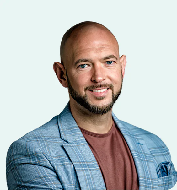 Smiling bald man with a beard wearing a light blue plaid blazer and a brown shirt against a light background.
